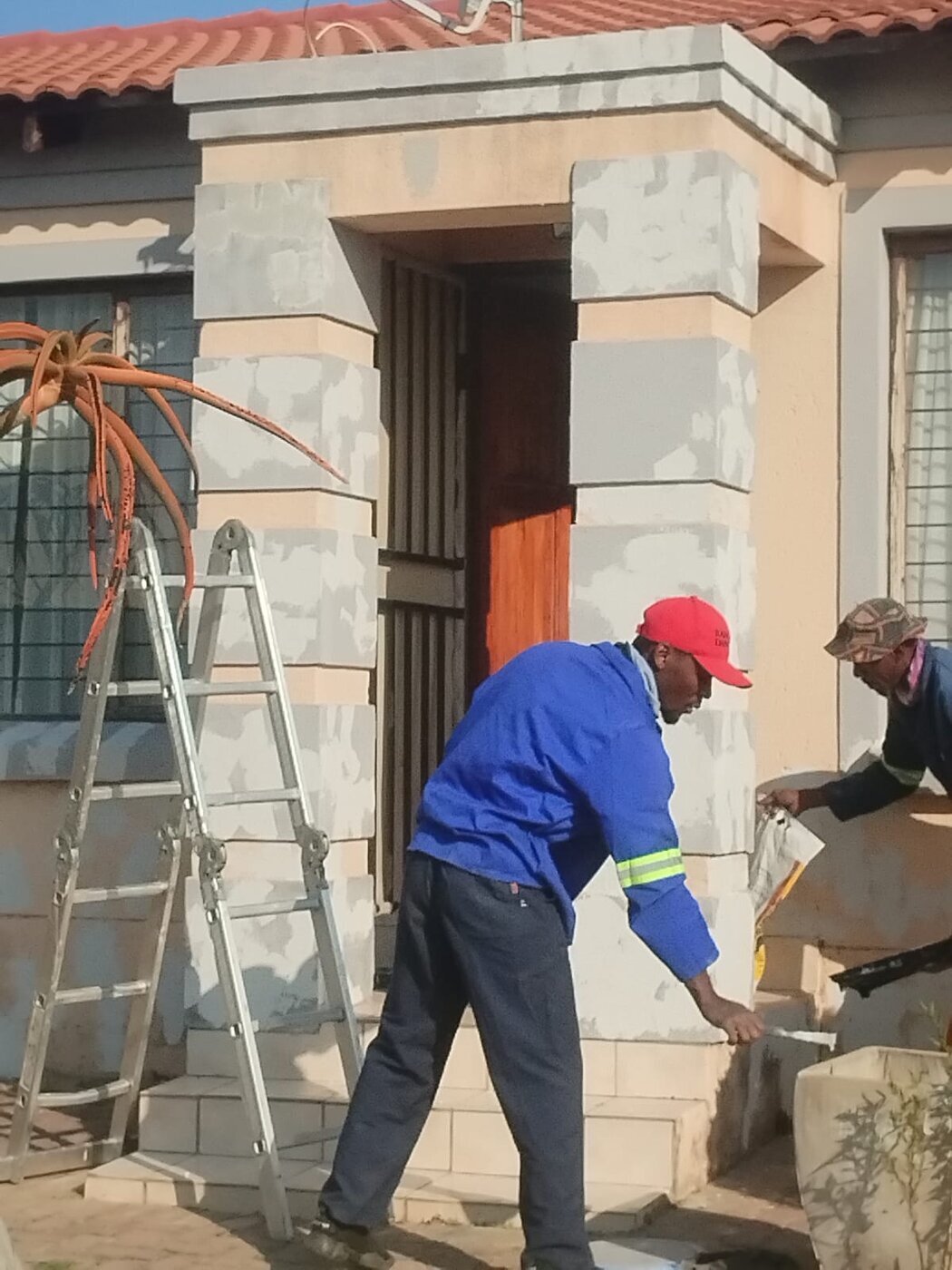 Two Thapelosego workers preparing exterior entrance pillars for painting.